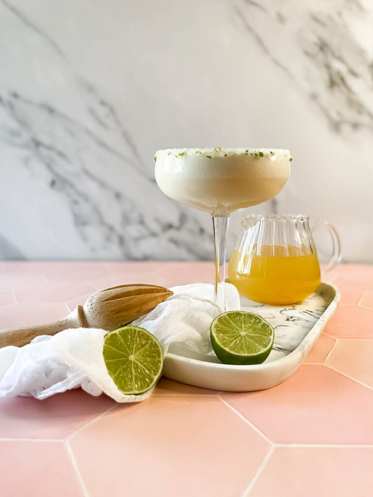 pineapple coconut margarita glass on a marble tray, with ice and lime on the foreground, a white napkin, a wooden juicer and a jug of juice and tequila bottle on the background