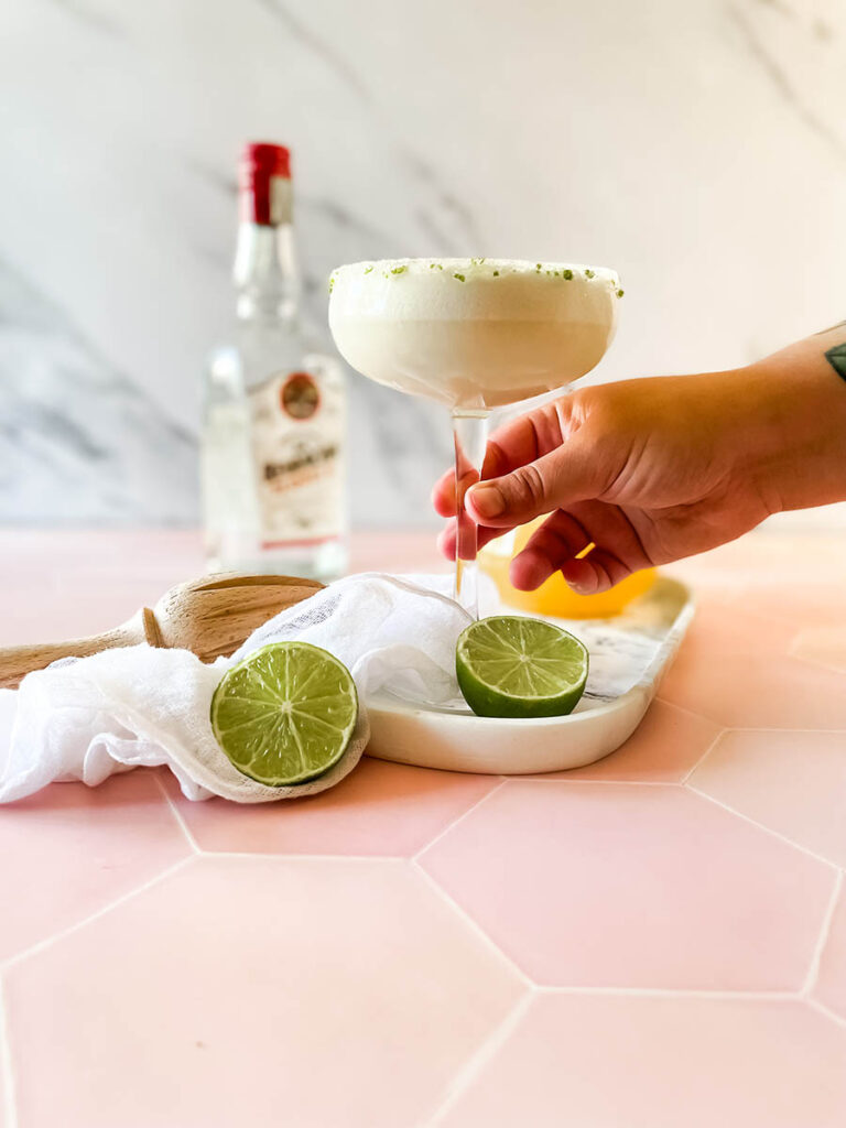 pineapple coconut margarita glass being held by a hand on a marble tray, with ice and lime on the foreground, a white napkin, a wooden juicer and a jug of juice and tequila bottle on the background