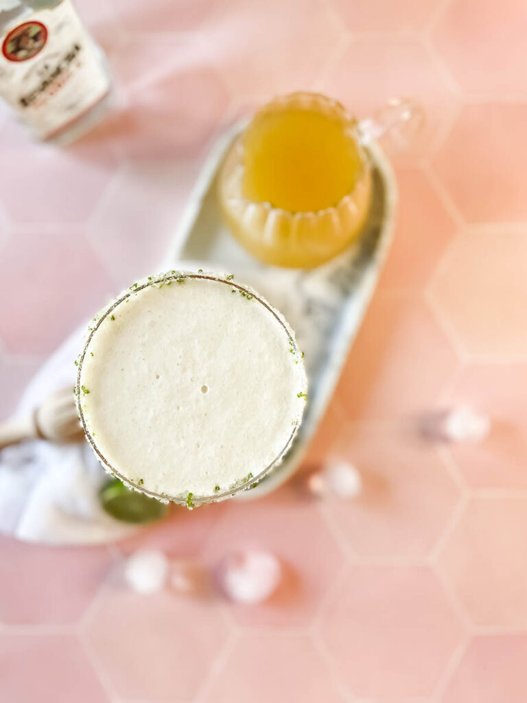 pineapple coconut margarita glass from above on a marble tray, with ice and lime on the foreground, a white napkin, a wooden juicer and a jug of juice and tequila bottle on the background