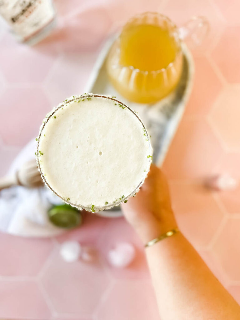 pineapple coconut margarita glass from above with a hand holding the glass, on a marble tray, with ice and lime on the foreground, a white napkin, a wooden juicer and a jug of juice and tequila bottle on the background