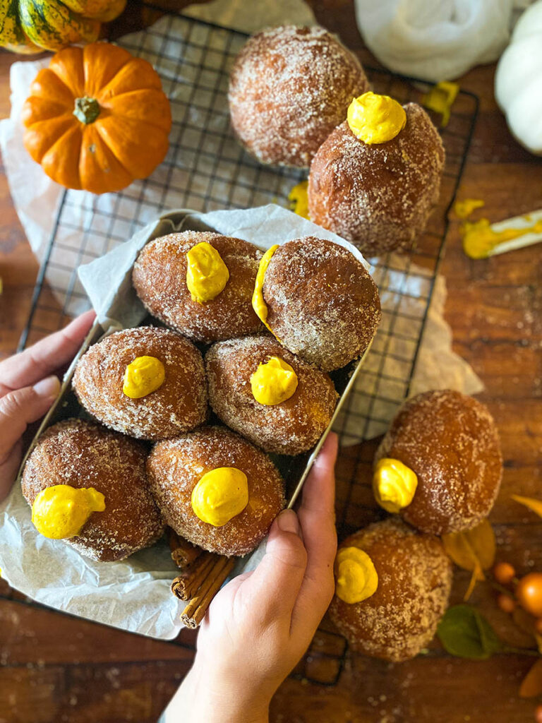 The Best Pumpkin Cheesecake Donuts: Perfect Fall Treat 1 pumpkin cheesecake donut recipe, donuts seen from above in a metal container being held by two hands, over a cooling rack