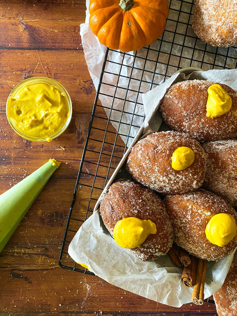 The Best Pumpkin Cheesecake Donuts: Perfect Fall Treat 8 pumpkin cheesecake donut recipe, donuts seen from above in a metal container over a cooling rack, with a piping bag filled with pumpkin cheesecase on the side