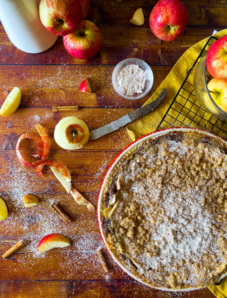 Delicious Apple Pie With Graham Cracker Crust 3 Overhead view of a freshly baked apple pie with a graham cracker crumb topping, dusted with powdered sugar. Surrounding the pie are whole and sliced apples, a bowl of flour, a knife, and cinnamon sticks scattered on a wooden surface, creating a cozy autumnal scene.