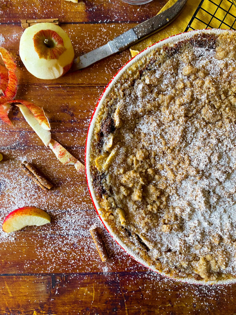 Delicious Apple Pie With Graham Cracker Crust 6 Overhead view of a freshly baked apple pie with a graham cracker crumb topping, dusted with powdered sugar. Surrounding the pie are whole and sliced apples, a bowl of flour, a knife, and cinnamon sticks scattered on a wooden surface, creating a cozy autumnal scene.
