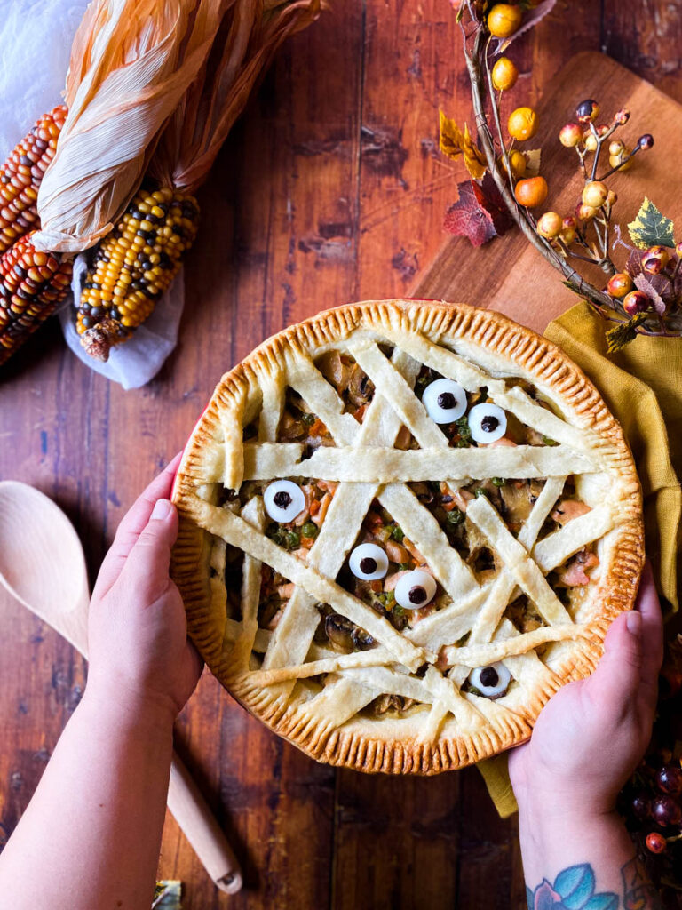 Easy Mummy pot Pie - Delicious Halloween Main Dish 4 hands holding mummy pot pie on a wooden background, corn, spoon and leaves around
