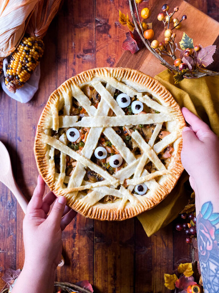 Easy Mummy pot Pie - Delicious Halloween Main Dish 2 hands holding mummy pot pie on a wooden background, corn, spoon and leaves around