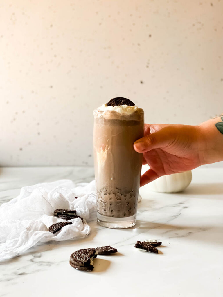 Close-up image of a glass filled with creamy Oreo Milk Tea. The drink is topped with whipped cream and garnished with crushed Oreo cookies. 