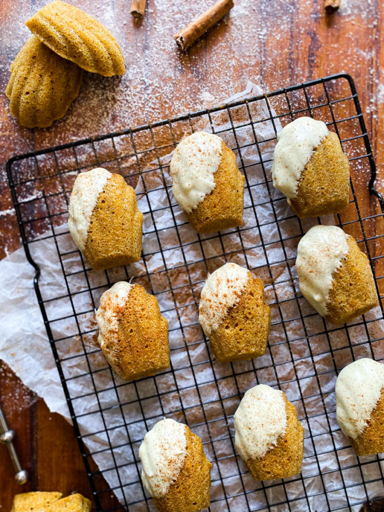 Delicious Pumpkin Madeleines With White Chocolate 6 close up of pumpkin madeleines on a cooling rack on a wooden background, from above