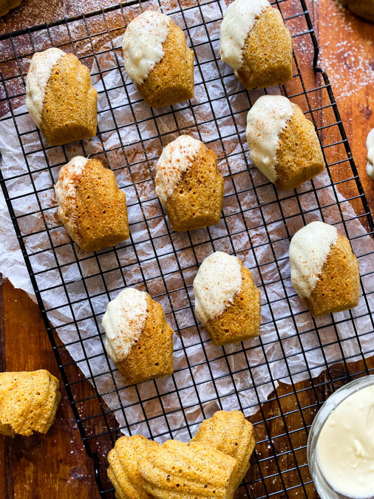 Delicious Pumpkin Madeleines With White Chocolate 1 close up pumpkin madeleines on a cooling rack on a wooden background, more madeleines on sides