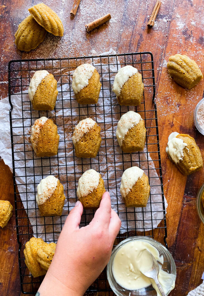Delicious Pumpkin Madeleines With White Chocolate 2 hand reaching for a pumpkin madeleine on a cooling rack on a wooden background