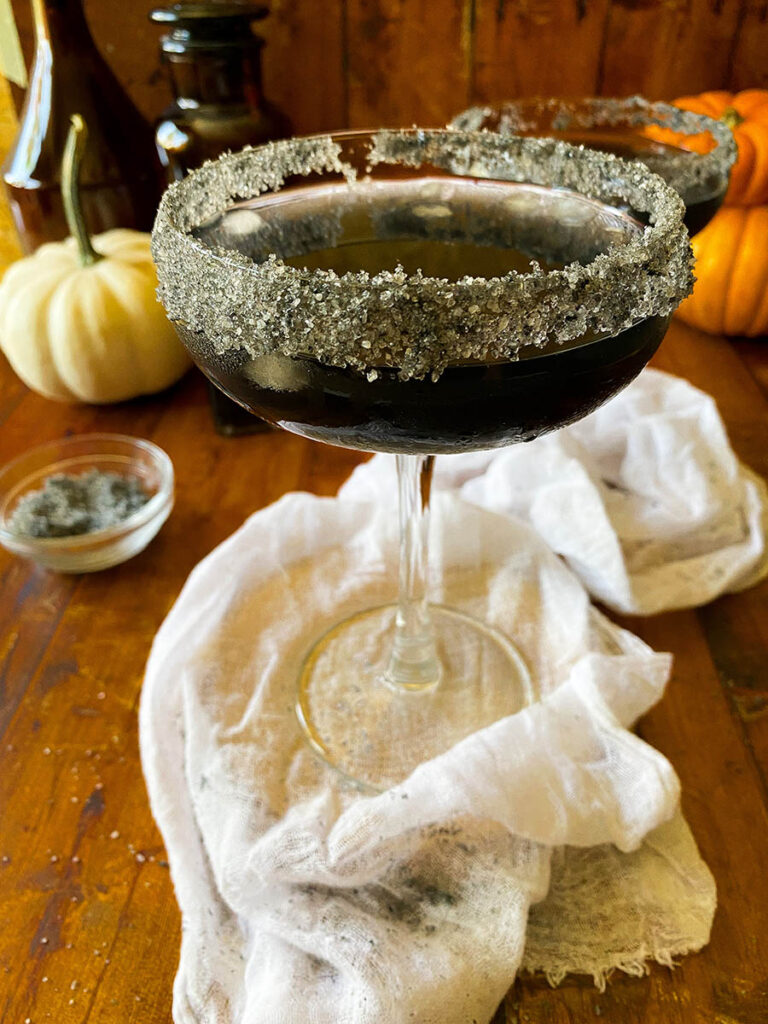 Scary Good Halloween Margarita Recipe 7 black halloween margaritas with black salt rims and a white napkin, with pumpkins on the background
