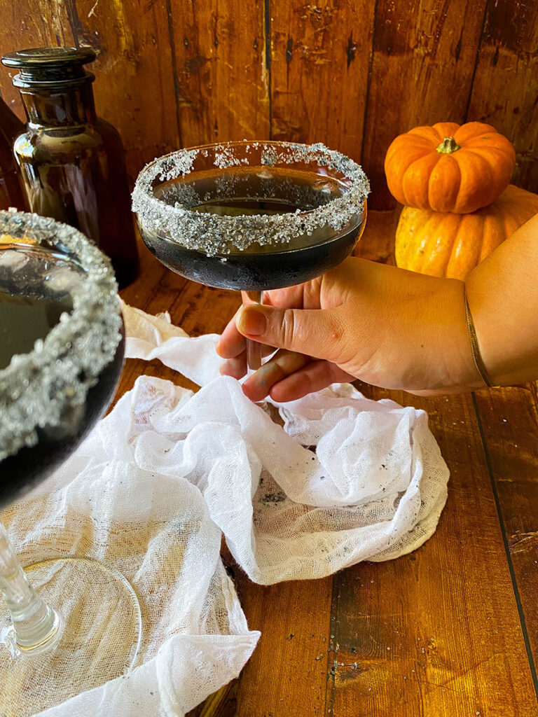 Scary Good Halloween Margarita Recipe 8 black halloween margaritas with black salt rims and a white napkin, with pumpkins on the background, with one of the glasses being held by a hand