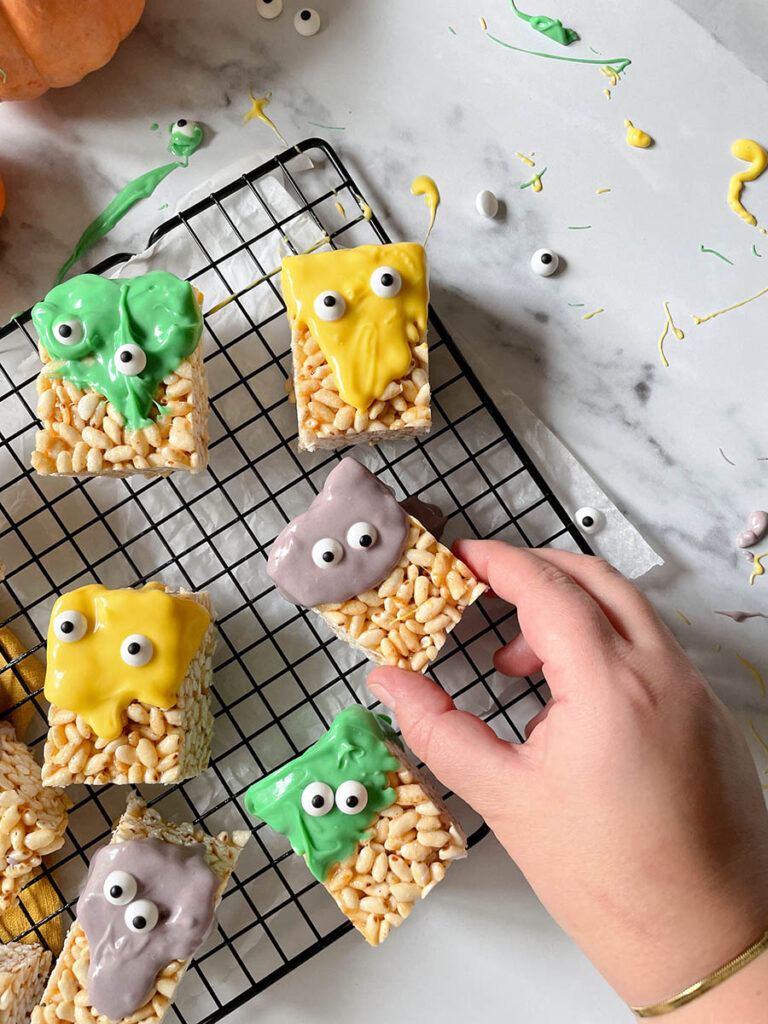 MM Rice Krispie Treats - The Delicious Monster Mash Treats 2 MM rice krispie treats on a cooling rack, being grabbed by a hand with pumpkins scattered around