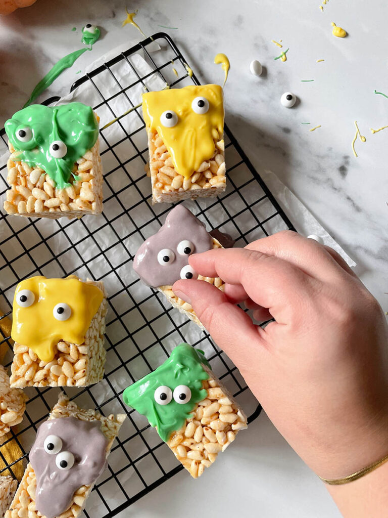 MM Rice Krispie Treats - The Delicious Monster Mash Treats 3 MM rice krispie treats on a cooling rack with a hand placing the eyes on and with pumpkins scattered around