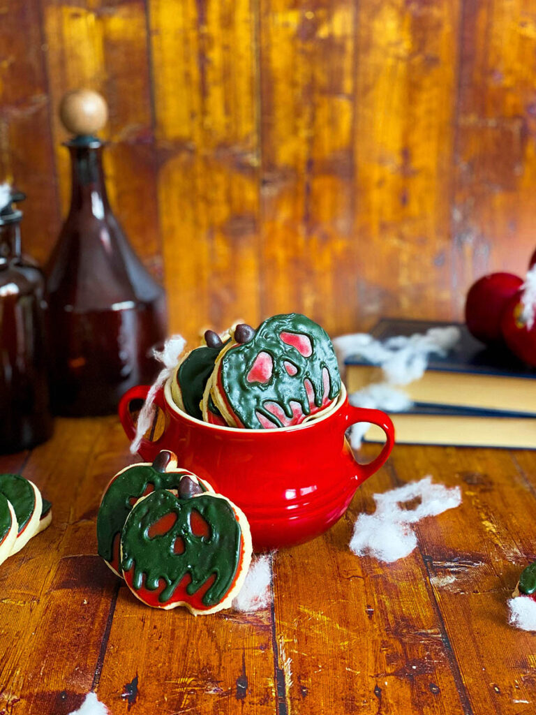 Delicious & Scary But Easy Poison Apple Cookies, Snow White's Inspired 2 poison apple cookie in a red cauldron with some spiderwebs scattered across, some poison bottles and books