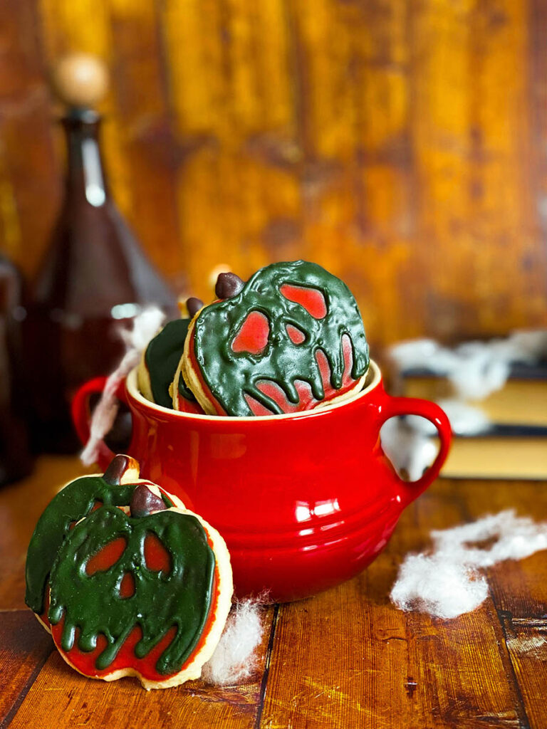Delicious & Scary But Easy Poison Apple Cookies, Snow White's Inspired 7 poison apple cookie in a red cauldron with some spiderwebs scattered across, some poison bottles and books