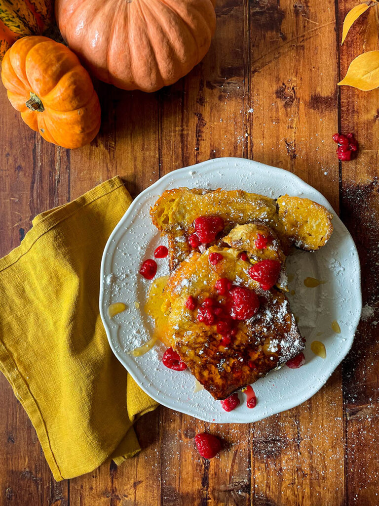 Delicious Pumpkin Brioche French Toast Recipe 3 pumpkin brioche french toast from above with honey and raspberries on a white plate with icing sugar, pumpkins and foliage on the background