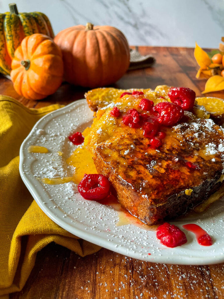 Delicious Pumpkin Brioche French Toast Recipe 5 pumpkin brioche french toast with honey and raspberries on a white plate with icing sugar, pumpkins and foliage on the background