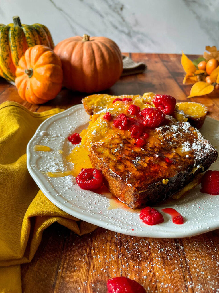 Delicious Pumpkin Brioche French Toast Recipe 9 pumpkin brioche french toast with honey and raspberries on a white plate with icing sugar, pumpkins and foliage on the background