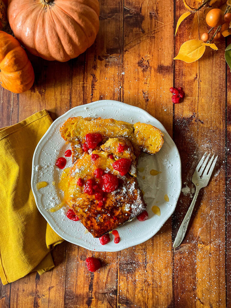 Delicious Pumpkin Brioche French Toast Recipe 6 pumpkin brioche french toast with honey and raspberries on a white plate with icing sugar, pumpkins and foliage on the background