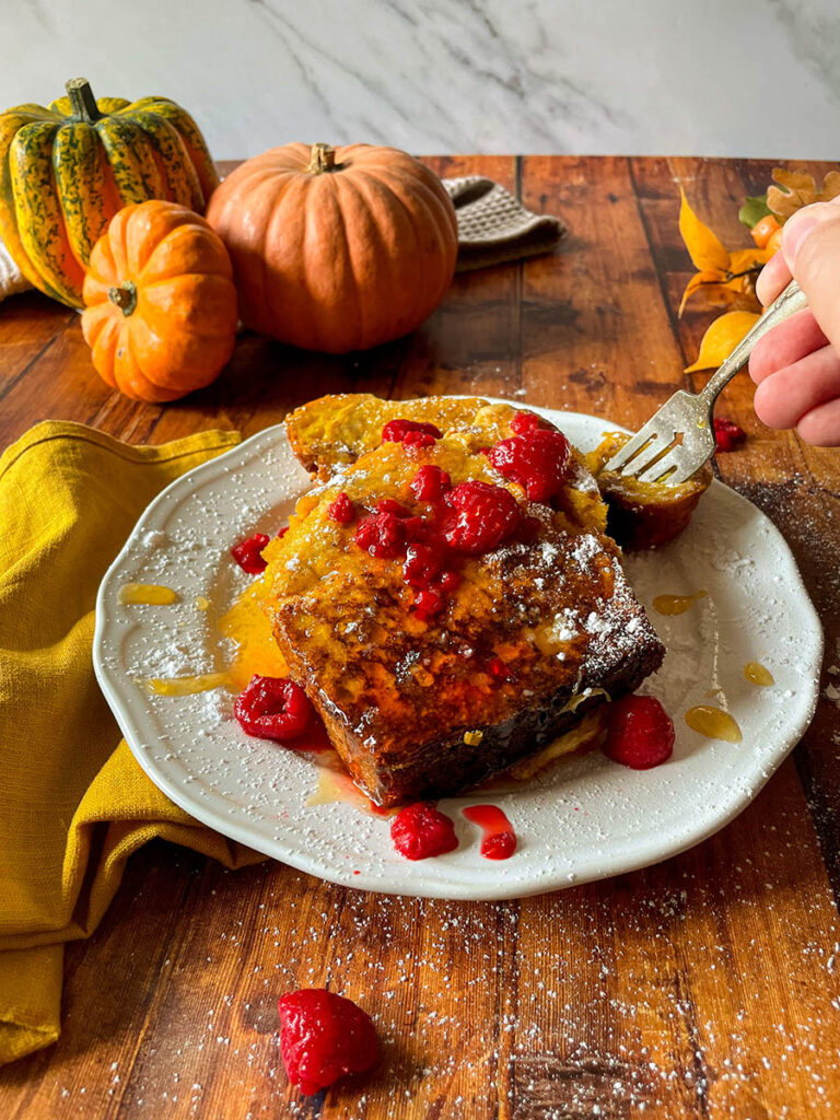 Delicious Pumpkin Brioche French Toast Recipe 11 pumpkin brioche french toast with honey and raspberries on a white plate with icing sugar, pumpkins and foliage on the background