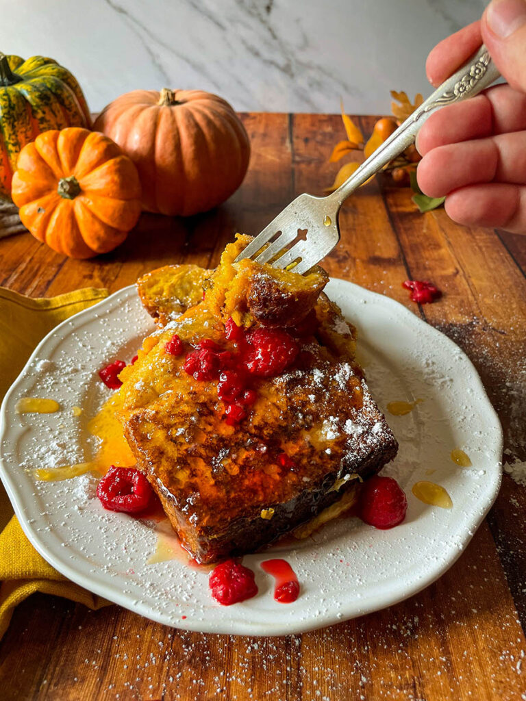Delicious Pumpkin Brioche French Toast Recipe 1 pumpkin brioche french toast with honey and raspberries on a white plate with icing sugar, pumpkins and foliage on the background