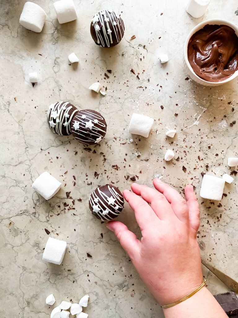 Baileys hot chocolate bombs over a marble countertop, with chocolate shavings, marshmallows and extra ganache in a small pot