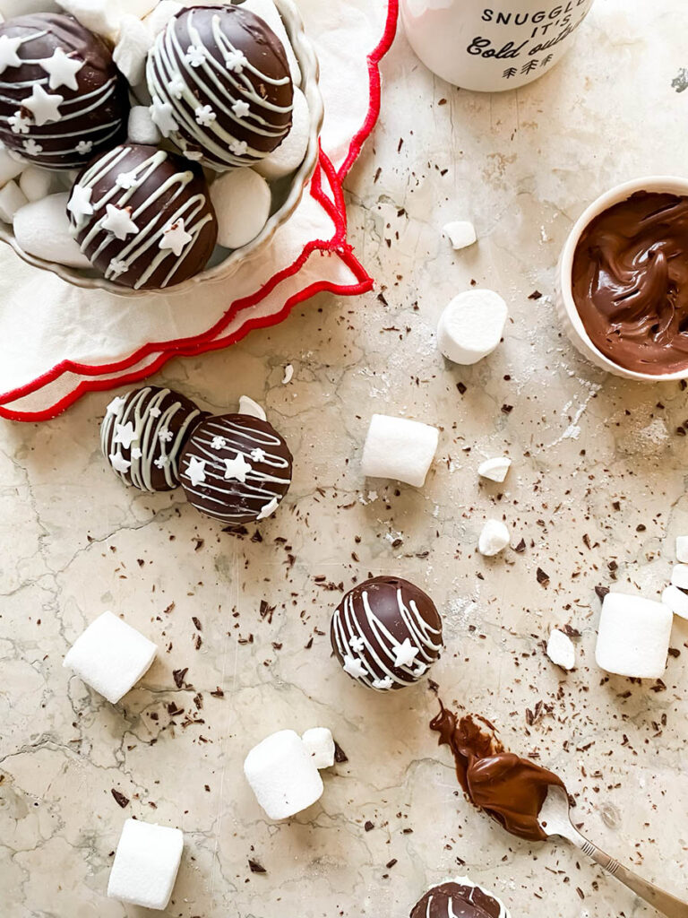 Baileys hot chocolate bombs over a marble countertop, with chocolate shavings, marshmallows and extra ganache in a small pot