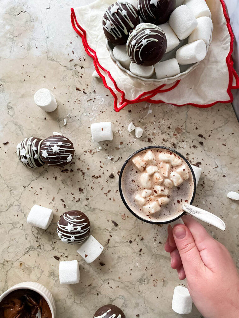 Baileys hot chocolate bombs over a marble countertop, with chocolate shavings, marshmallows and extra ganache in a small pot, plus a mug of hot chocolate ready to enjoy