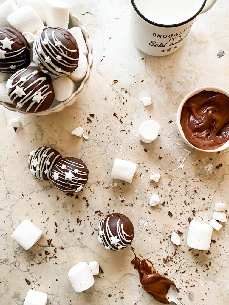 Baileys hot chocolate bombs over a marble countertop, with chocolate shavings, marshmallows and extra ganache in a small pot