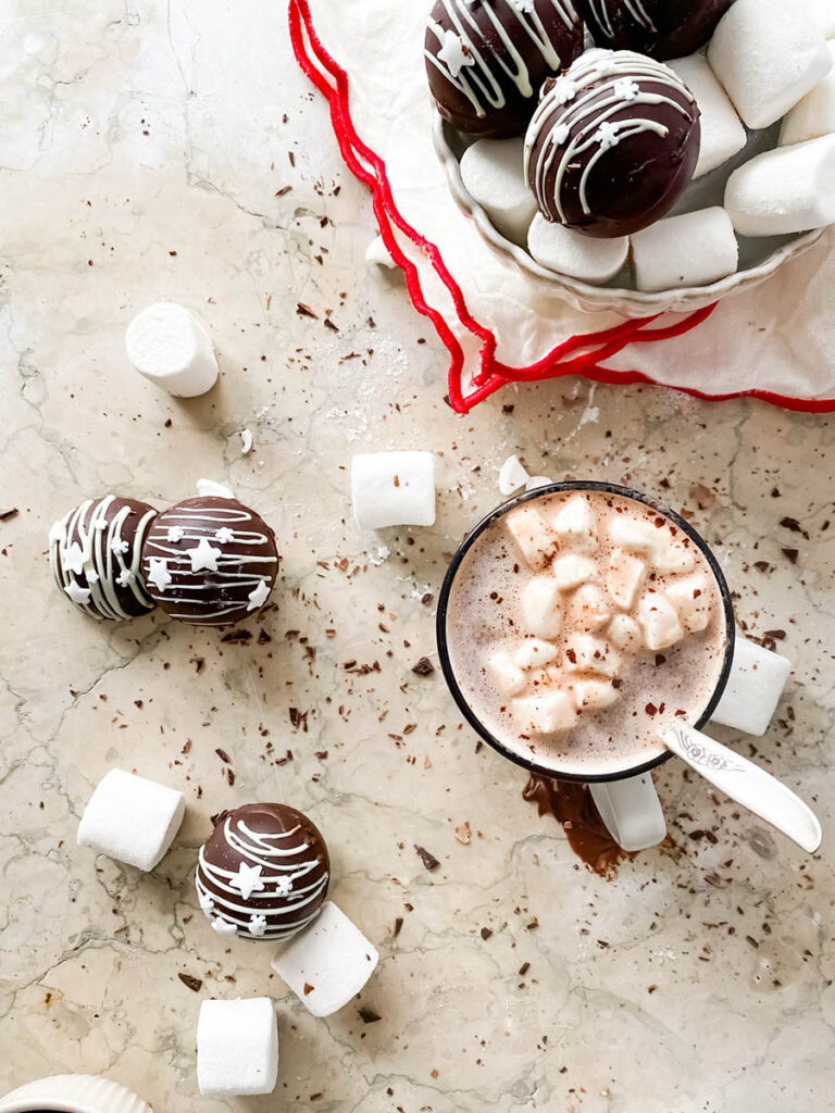 Baileys hot chocolate bombs over a marble countertop, with chocolate shavings, marshmallows and extra ganache in a small pot, and a mug of ready made hot chocolate ready to be enjoyed