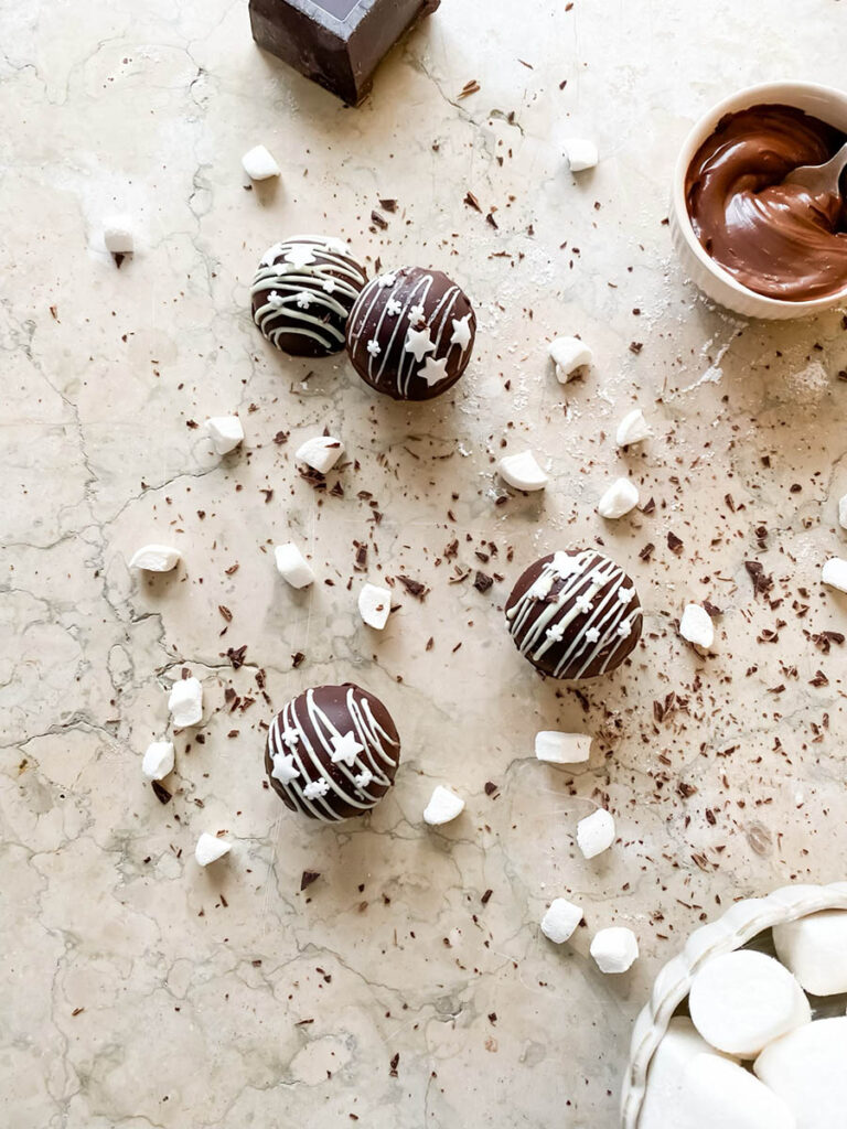 Baileys hot chocolate bombs over a marble countertop, with chocolate shavings, marshmallows and extra ganache in a small pot