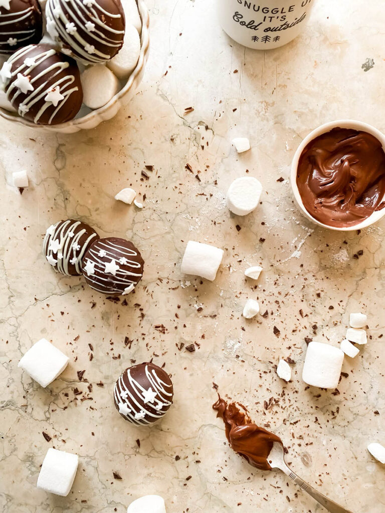 Baileys hot chocolate bombs over a marble countertop, with chocolate shavings, marshmallows and extra ganache in a small pot