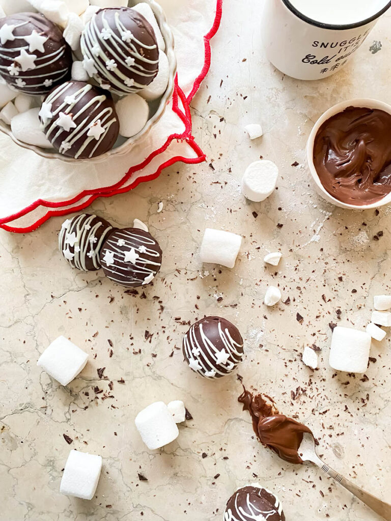 Baileys hot chocolate bombs over a marble countertop, with chocolate shavings, marshmallows and extra ganache in a small pot