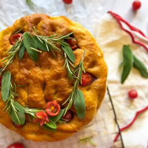 Christmas Focaccia: The Most Delicious Bread With A Festive Twist 3 close up christmas focaccia on parchment paper on top of a wire rack, tomatoes, sage and rosemary around, towels, ribbons and fir on marble background