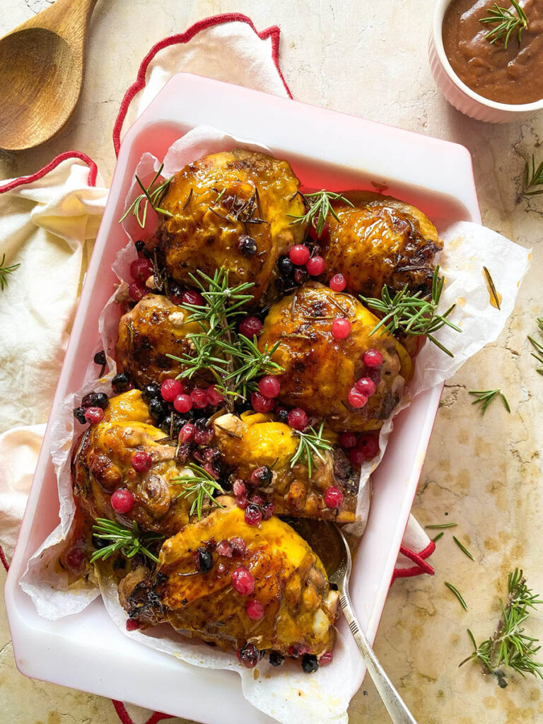 cranberry rosemary chicken in a baking tray over a red rimmed napkin, with a spoon and some fresh rosemary scattered around