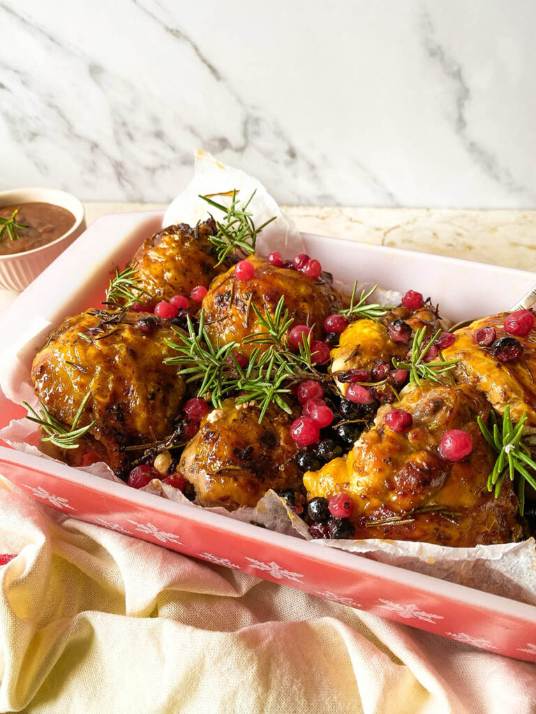 cranberry rosemary chicken in a baking tray over a red rimmed napkin, with a spoon and some fresh rosemary scattered around