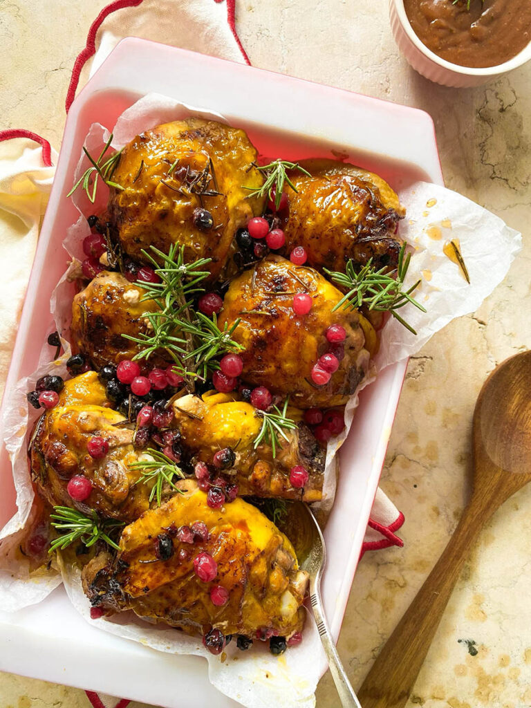 cranberry rosemary chicken in a baking tray over a red rimmed napkin, with a spoon and some fresh rosemary scattered around