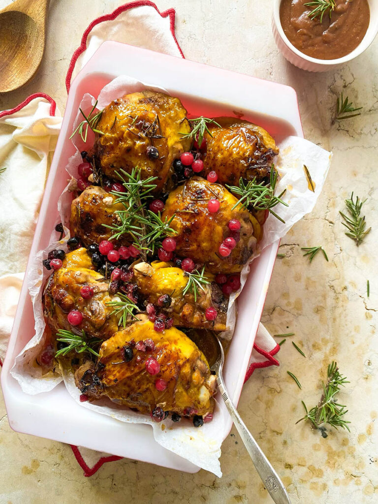 cranberry rosemary chicken in a baking tray over a red rimmed napkin, with a spoon and some fresh rosemary scattered around