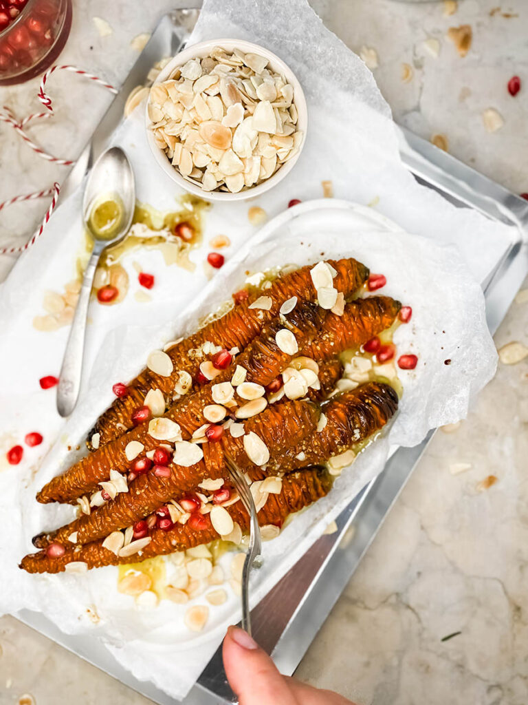 hassleback carrots on a silver tray with shaved almonds and pomegranate seeds over a marble top 