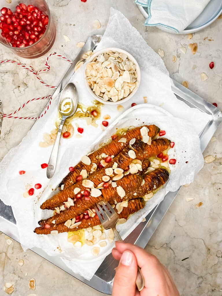 hassleback carrots on a silver tray with shaved almonds and pomegranate seeds over a marble top 