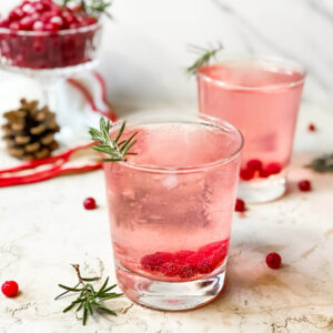 Super Mistletoe Kiss Cocktail Recipe for a Jolly Holiday Season 5 two glasses of mistletoe kiss, in the background cranberries, white and red towel, pinecones, around cranberries and rosemary, on marble background