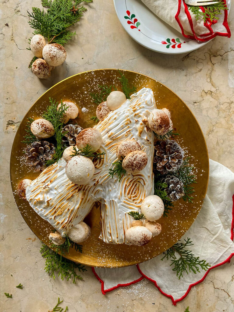 salted caramel yule log on a gold plate with meringue mushrooms, pine cones and green leaves to decorate