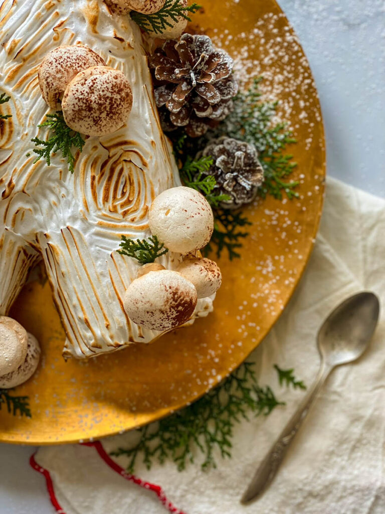 salted caramel yule log on a gold plate with meringue mushrooms, pine cones and green leaves to decorate
