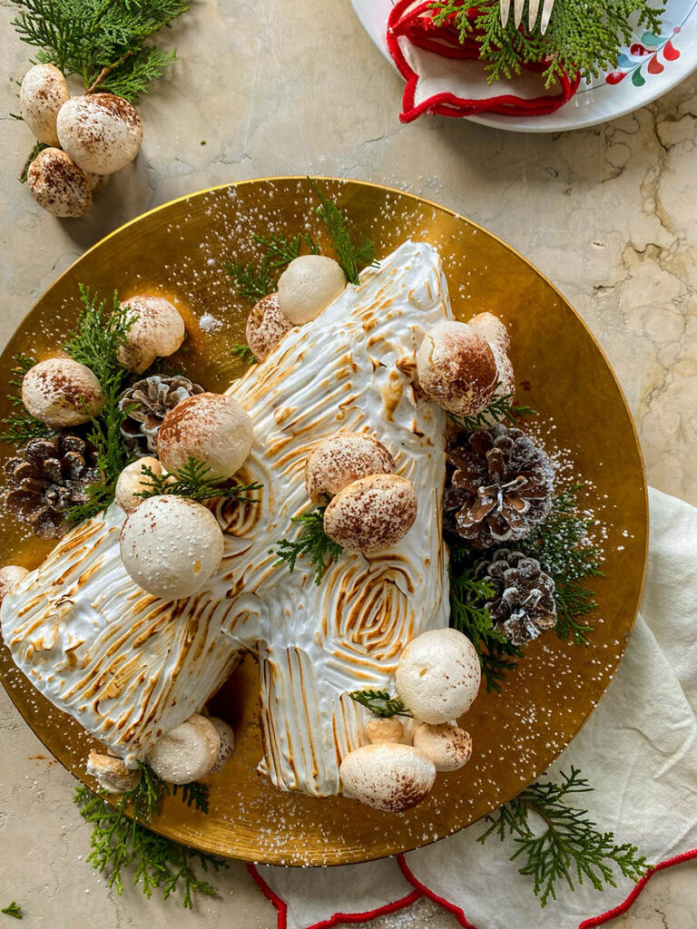 salted caramel yule log on a gold plate with meringue mushrooms, pine cones and green leaves to decorate