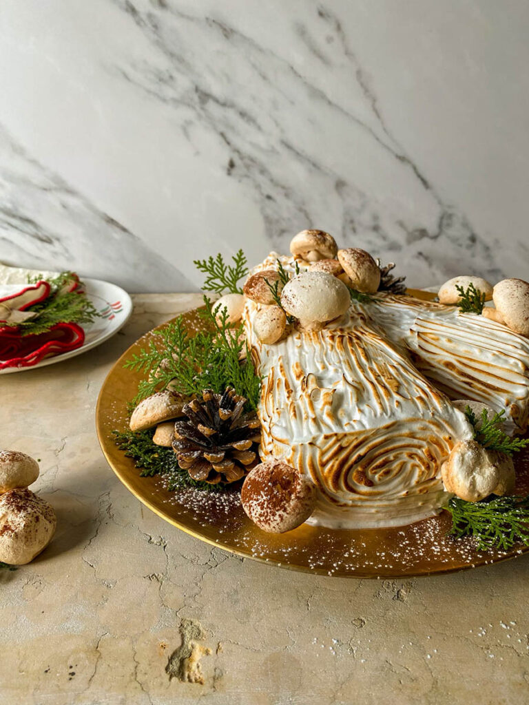 salted caramel yule log on a gold plate with meringue mushrooms, pine cones and green leaves to decorate