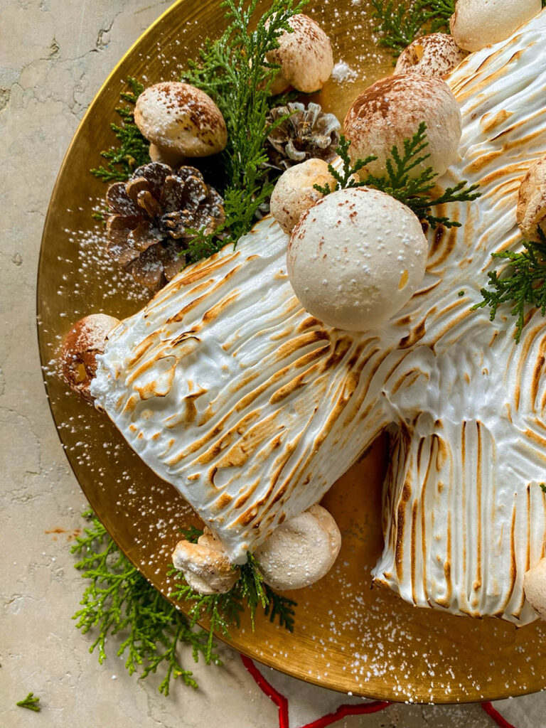 salted caramel yule log on a gold plate with meringue mushrooms, pine cones and green leaves to decorate