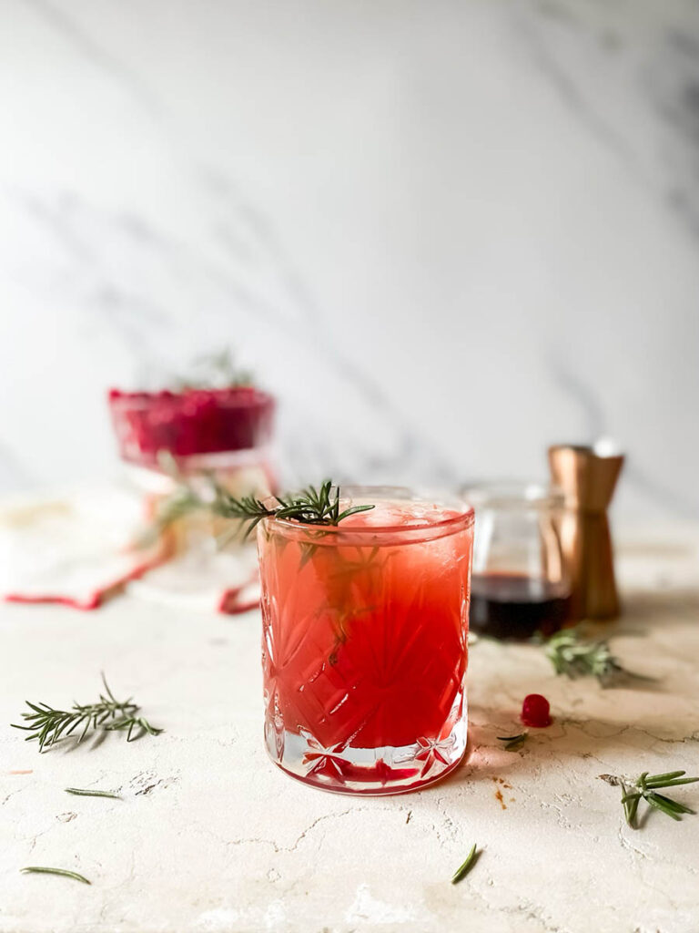 santa's tipsy spritzer cocktail in a whisky glass with rosemary as garnish, a measuring cup and cranberries in a glass coupe in the background