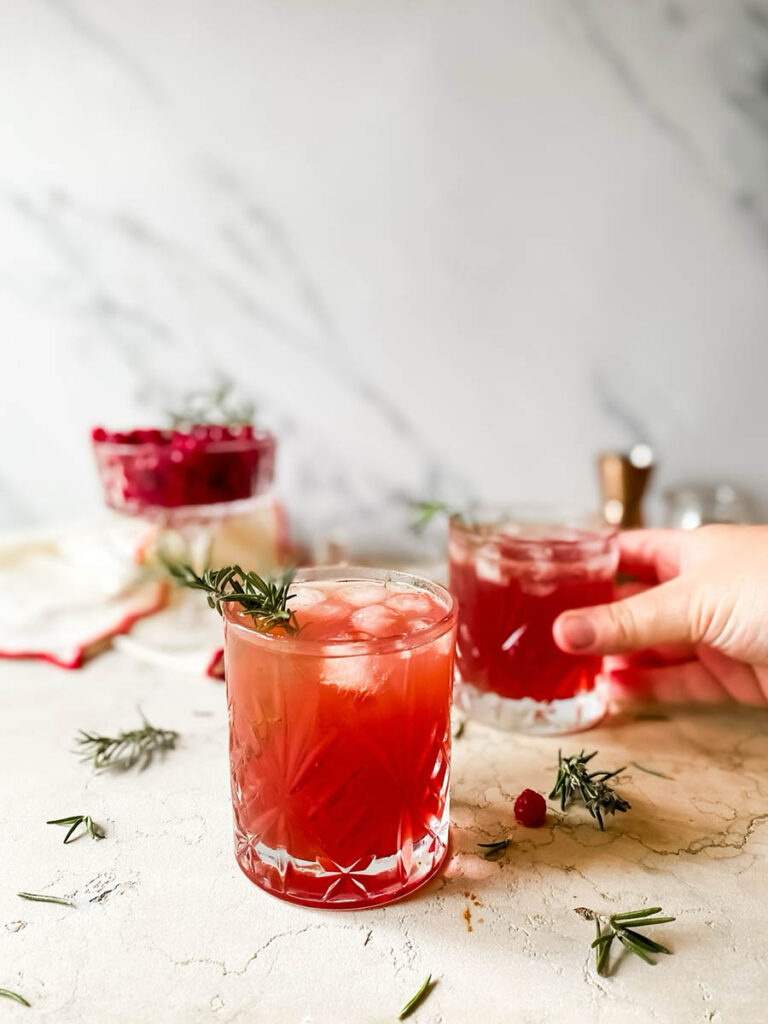 santa's tipsy spritzer cocktail in a whisky glass with rosemary as garnish, a measuring cup and cranberries in a glass coupe in the background