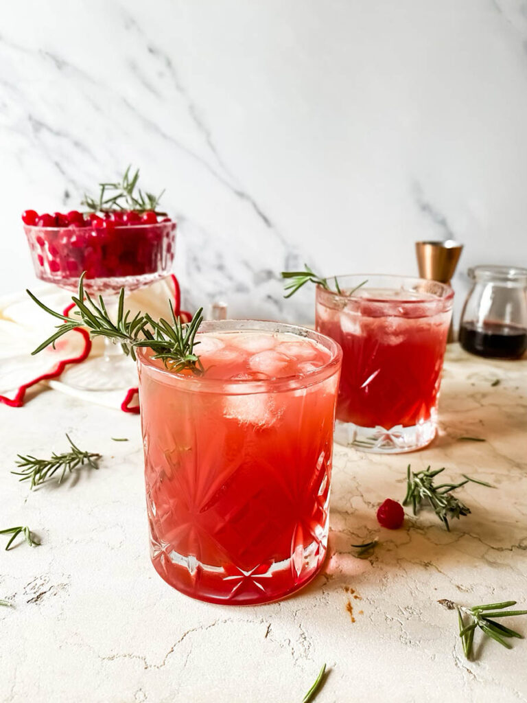 santa's tipsy spritzer cocktail in a whisky glass with rosemary as garnish, a measuring cup and cranberries in a glass coupe in the background